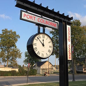 36-inch diameter double-faced Suspended Bracket Clock mounted below a sign for Port Colborne Transit Station, in Port Colborne, Ontario, Canada. Background of blue sky.